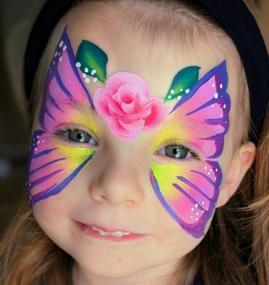 Mädchen mit buntem Schmetterlingsgesichtsbemalung und einer rosa Blume auf der Stirn.